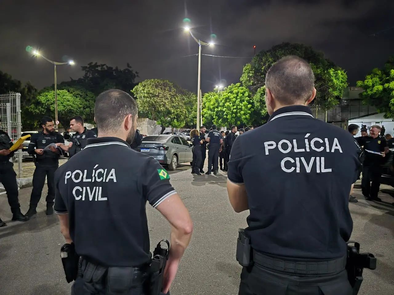 Polícia Civil realizando operação noturna com agentes uniformizados na rua, com árvores e iluminação pública ao fundo, durante a noite.