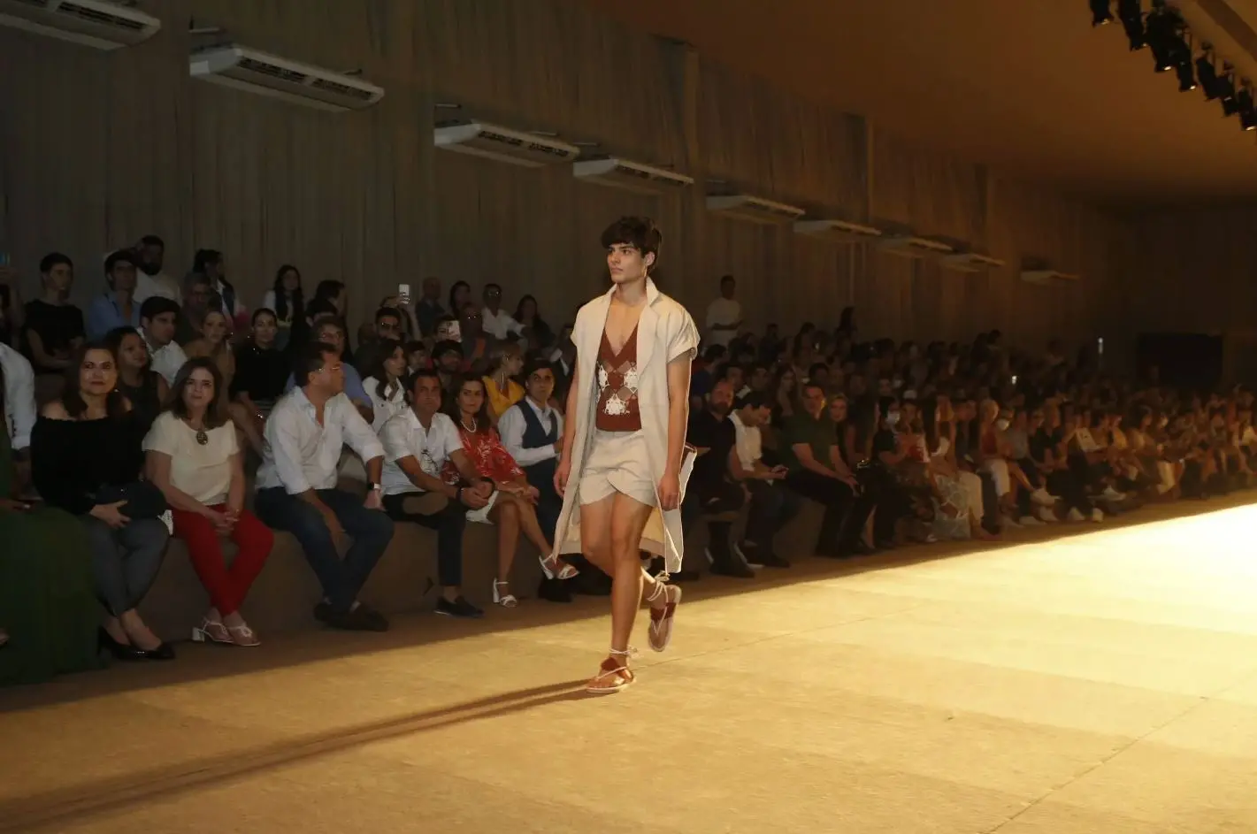 Pessoa caminha por uma passarela iluminada durante um desfile de moda, usando roupa clara com top marrom estampado e sandálias. Ao fundo, uma plateia sentada acompanha o evento em um grande auditório.