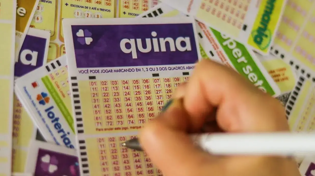 Mão segura uma caneta e marca números em um volante da loteria Quina, com outros bilhetes de apostas ao fundo.