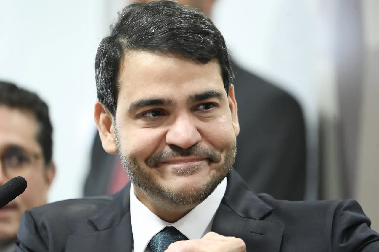 Pessoa vestindo terno escuro, camisa branca e gravata, sentada em ambiente interno, com microfone visível à esquerda e outras pessoas desfocadas ao fundo.