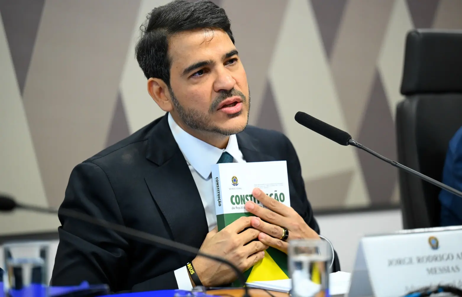 Jorge Messias é um homem jovem, branco, de barba rala e cabelo preto. Na foto, ele está sendo sabatinado no Senado e segura um exemplar da Constituição.