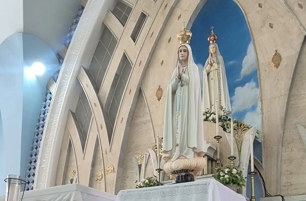 Duas estátuas de Nossa Senhora de Fátima posicionadas no altar de uma igreja. A imagem em primeiro plano veste manto branco com detalhes dourados e segura um terço, enquanto a segunda imagem, ao fundo, está emoldurada por um nicho azul que simula o céu com nuvens.