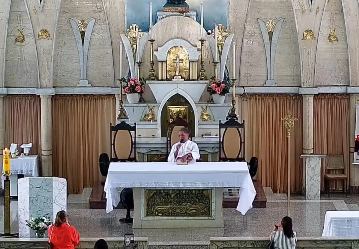 Padre celebra missa atrás de um altar coberto por uma toalha branca na Igreja de Fátima. O presbitério possui elementos em mármore e decorações douradas, com fiéis dispersos nos bancos em primeiro plano acompanhando a celebração.