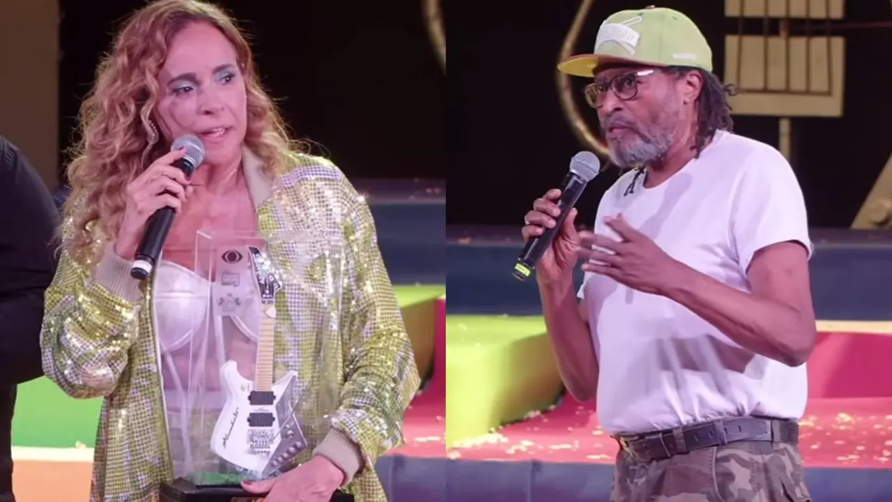 Daniela Mercury e Edson Gomes seguram microfones no palco, com a cantora segurando um troféu. Mercury veste uma jaqueta dourada brilhante, enquanto Gomes usa uma camiseta branca e boné verde.