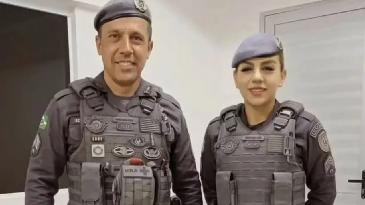 Duas pessoas lado a lado em ambiente interno, vestindo uniformes táticos cinza com coletes e boinas, diante de uma parede clara.