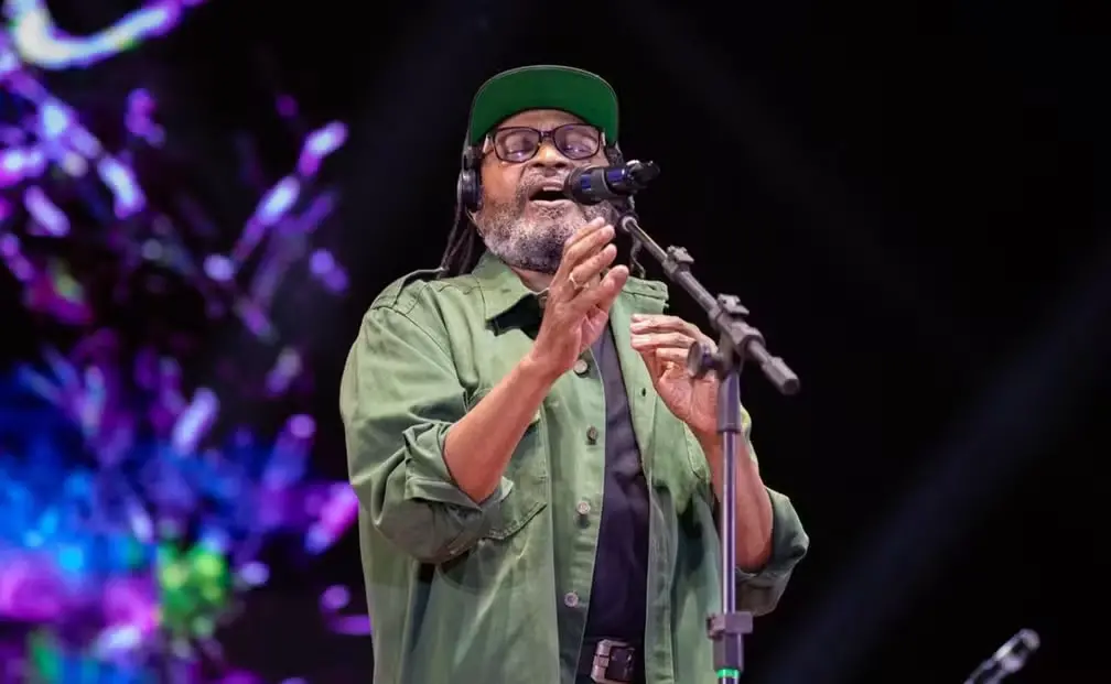 O cantor Edson Gomes se apresenta no palco, cantando expressivamente com as mãos próximas ao microfone e olhos fechados em um momento de entrega. Ele veste uma camisa verde sobreposta a uma camiseta preta e usa um boné verde, com luzes coloridas desfocadas ao fundo.