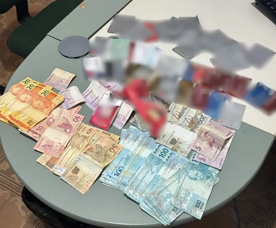 Imagem mostra uma mesa clara, em ambiente interno, com diversas cédulas de real espalhadas e organizadas em pequenos montes. Há notas de diferentes valores, incluindo 5, 10, 20, 50 e 100 reais. Sobre o dinheiro, aparecem cartões bancários e papéis parcialmente borrados para ocultar informações sensíveis. Ao fundo, é possível ver parte de uma cadeira e um teclado, indicando ambiente de trabalho ou escritório.