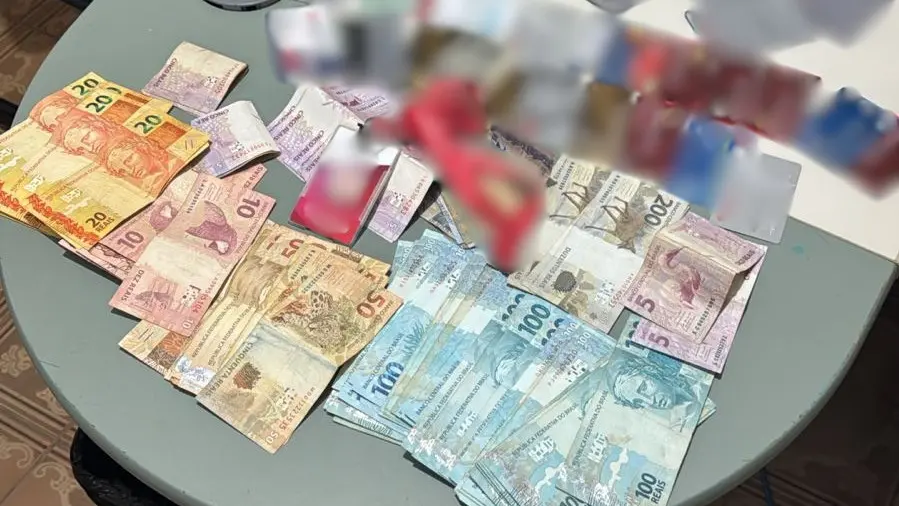 Imagem mostra uma mesa clara, em ambiente interno, com diversas cédulas de real espalhadas e organizadas em pequenos montes. Há notas de diferentes valores, incluindo 5, 10, 20, 50 e 100 reais. Sobre o dinheiro, aparecem cartões bancários e papéis parcialmente borrados para ocultar informações sensíveis. Ao fundo, é possível ver parte de uma cadeira e um teclado, indicando ambiente de trabalho ou escritório.