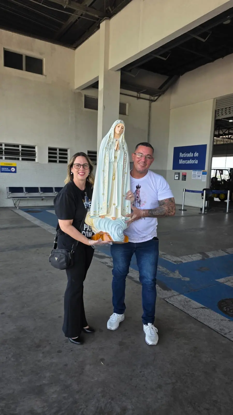 Um homem e uma mulher sorriem enquanto seguram juntos uma imagem de Nossa Senhora de Fátima em um pátio de cargas do aeroporto de Fortaleza. Eles estão em frente a uma placa que indica Retirada de Mercadoria, marcando a chegada da estátua sacra ao local.