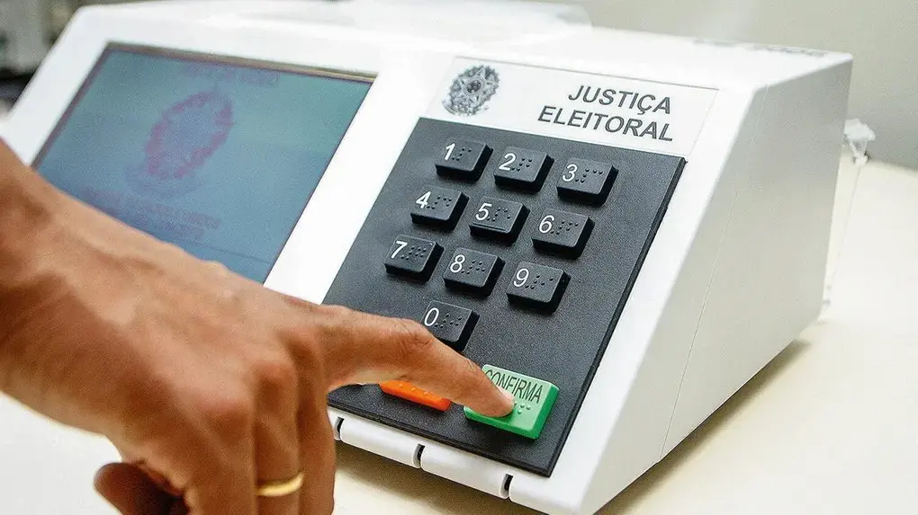 Pessoa pressiona o botão verde “CONFIRMA” de uma urna eletrônica da Justiça Eleitoral brasileira, com teclado numérico visível e tela ao lado, durante o processo de votação.