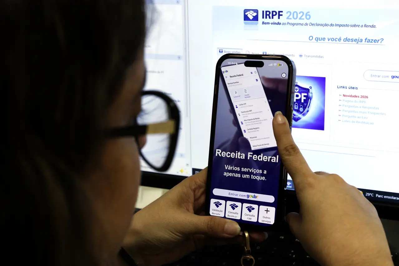 Uma pessoa segura um smartphone exibindo o aplicativo da Receita Federal à frente de um monitor que mostra o programa IRPF 2026. A imagem foca na interação digital para a declaração do imposto de renda.