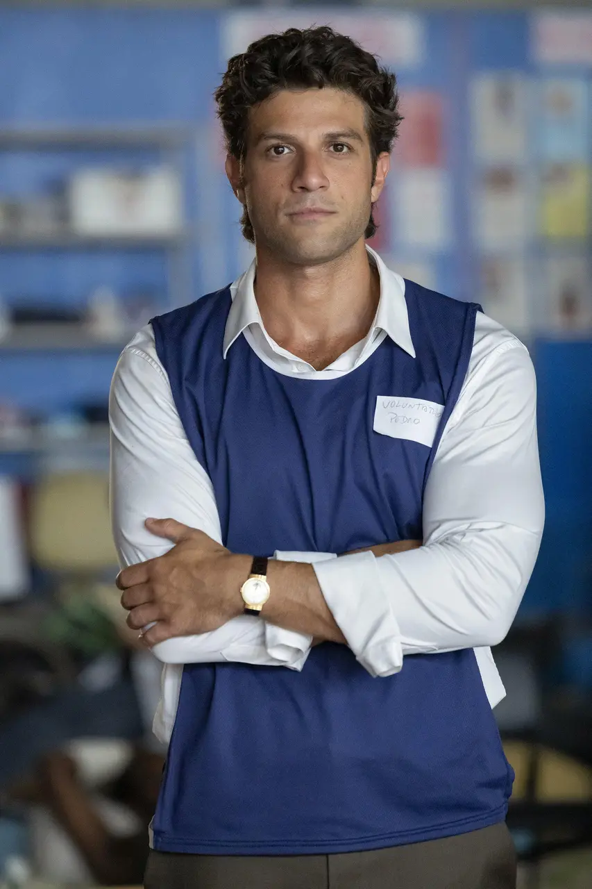 Pessoa em pé, com braços cruzados, vestindo camisa clara e colete azul com crachá escrito “Voluntário”, em ambiente interno com prateleiras desfocadas ao fundo.