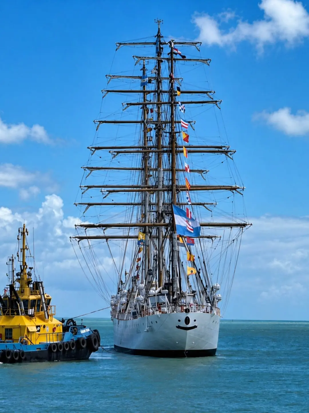 Fragata argentina branca com múltiplos mastros e bandeiras coloridas navega no oceano, acompanhado por um rebocador amarelo e azul ao seu lado. A embarcação destaca-se sob um céu azul com nuvens brancas, compondo uma cena marítima vibrante e imponente.