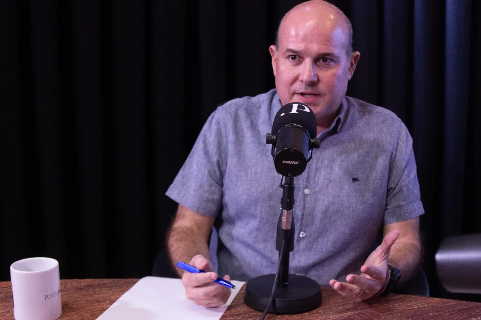 Homem calvo, usando camisa cinza de manga curta, fala em frente a um microfone durante gravação de podcast ou entrevista em estúdio. Ele está sentado à mesa de madeira, segurando uma caneta azul e gesticulando com a outra mão. Ao fundo, há cortinas pretas e iluminação em tom arroxeado. Uma caneca branca aparece no canto esquerdo da mesa.