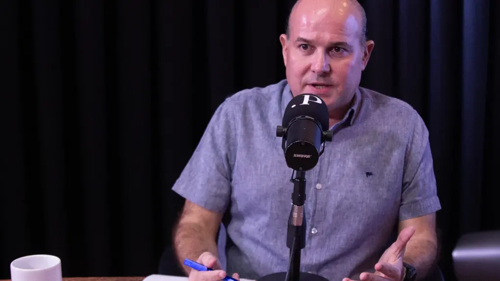 Homem calvo, usando camisa cinza de manga curta, fala em frente a um microfone durante gravação de podcast ou entrevista em estúdio. Ele está sentado à mesa de madeira, segurando uma caneta azul e gesticulando com a outra mão. Ao fundo, há cortinas pretas e iluminação em tom arroxeado. Uma caneca branca aparece no canto esquerdo da mesa.