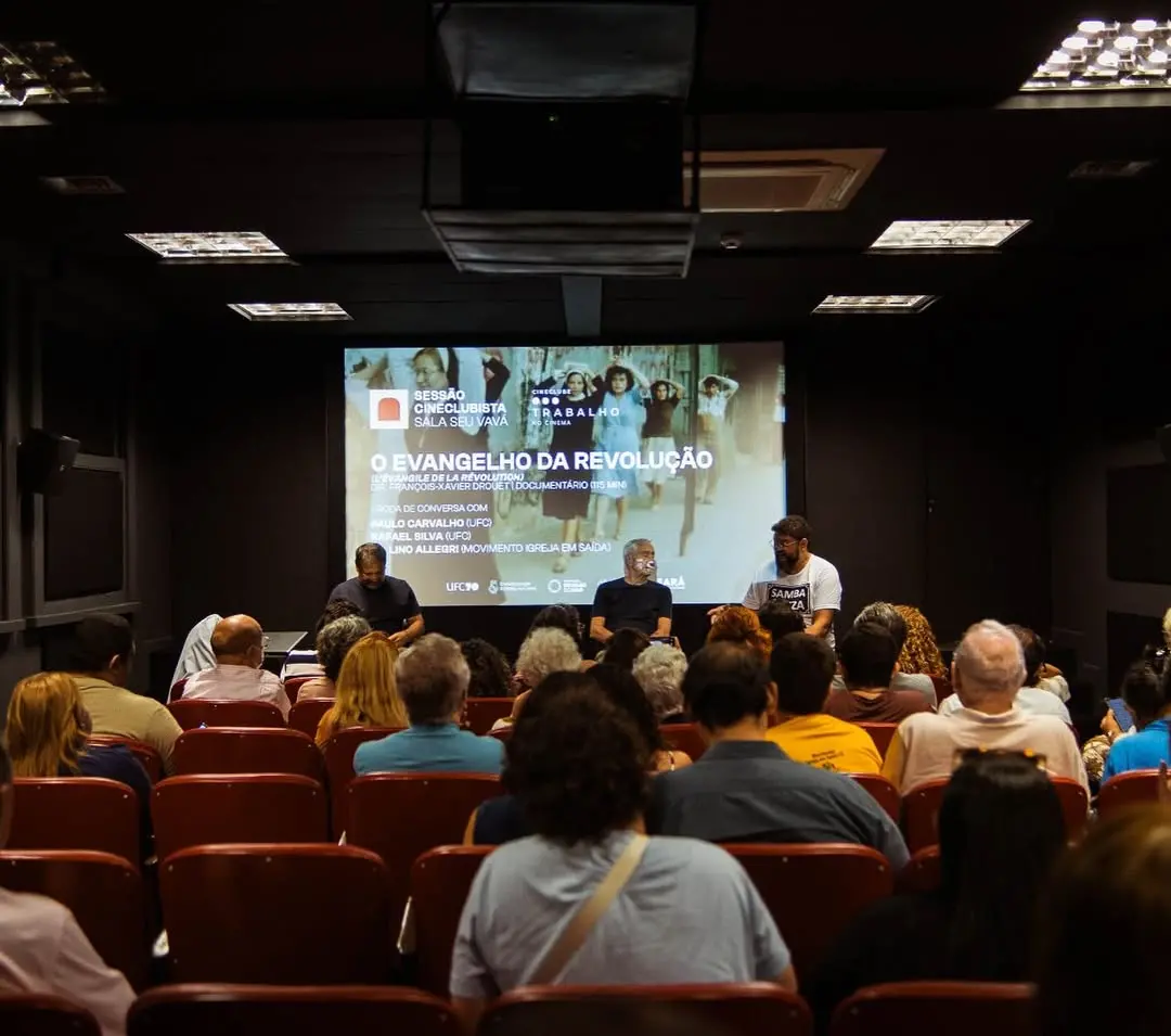 Três debatedores estão sentados à frente de uma plateia em um auditório escuro durante a sessão cineclubista do filme 'O Evangelho da Revolução'. A projeção ao fundo exibe o título do evento e informações sobre a roda de conversa, compondo um ambiente de engajamento cultural e debate.