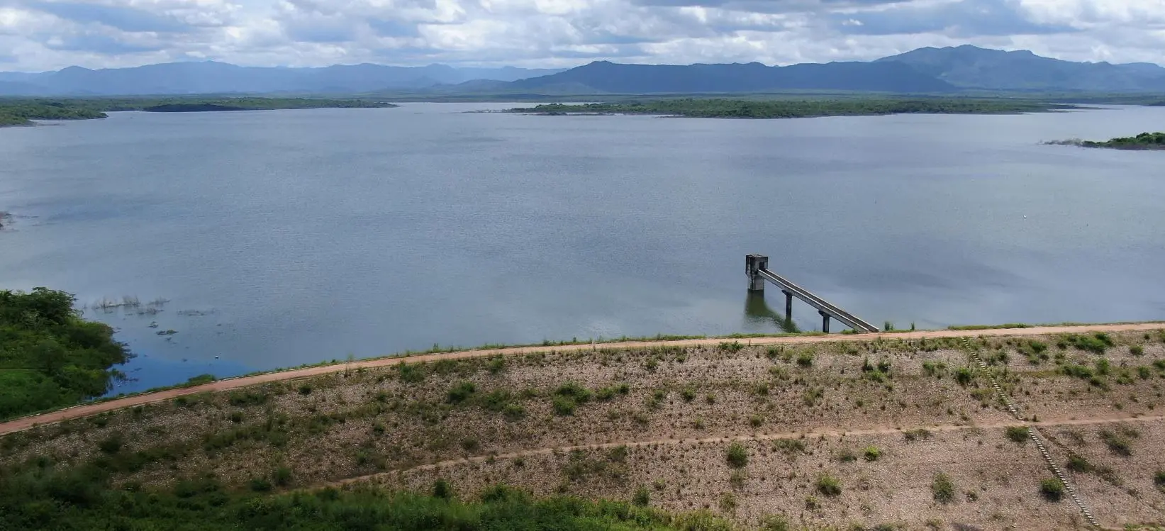 Imagem aérea de um vasto açude no Ceará, chamado Aracoiaba, barrado por uma grande parede de terra e um píer de monitoramento. Ao fundo, há morros e nuvens.