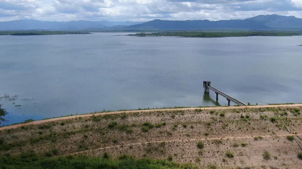 Imagem aérea de um vasto açude no Ceará, chamado Aracoiaba, barrado por uma grande parede de terra e um píer de monitoramento. Ao fundo, há morros e nuvens.