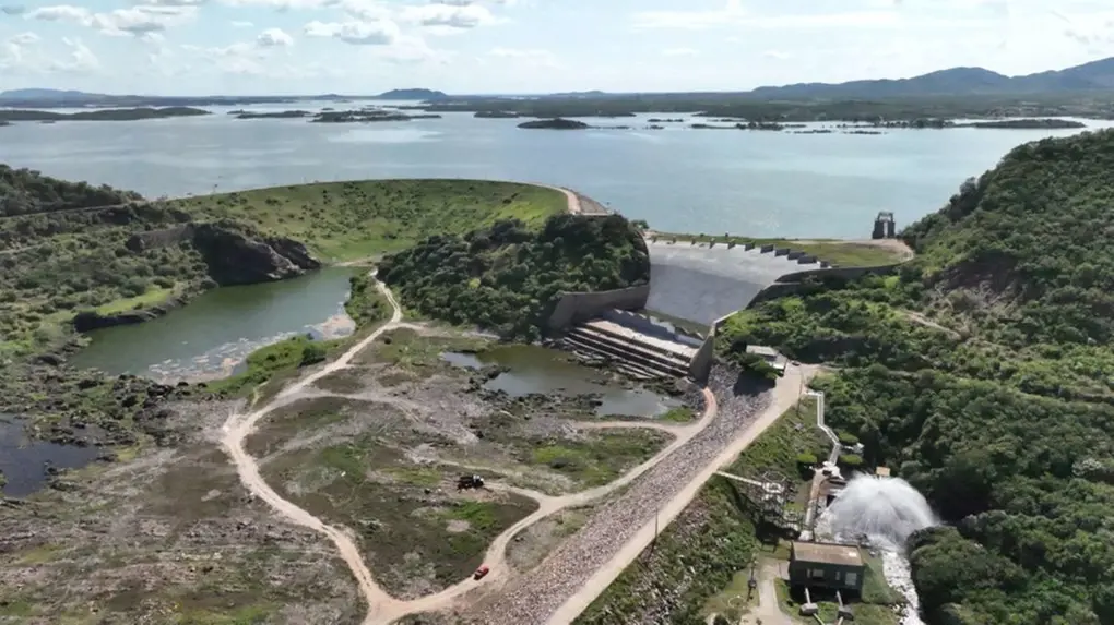 Vista aérea panorâmica de uma grande barragem, o Orós, cercada por montanhas e vegetação, com um vasto reservatório de água ao fundo sob um céu nublado. Na parte inferior direita, uma válvula de dispersão libera um forte jato de água branca, enquanto estradas de terra serpenteiam pelo terreno árido próximo à estrutura.