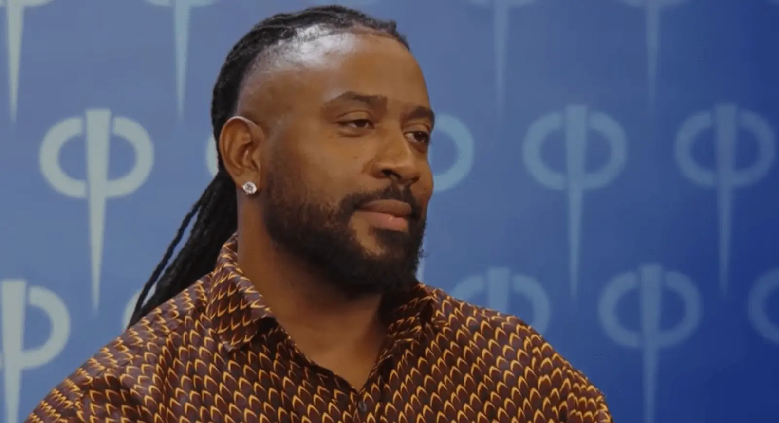 Homem com cabelo em tranças longas presas para trás e barba cheia aparece em close, usando uma camisa estampada em tons de marrom e dourado. Ele veste brincos discretos e olha levemente para o lado, com expressão séria. Ao fundo, há um painel azul com padrões gráficos repetidos.