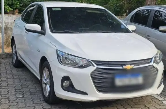 Automóvel Chevrolet branco, modelo compacto sedã, fotografado de frente em estacionamento ao ar livre com árvores ao fundo.