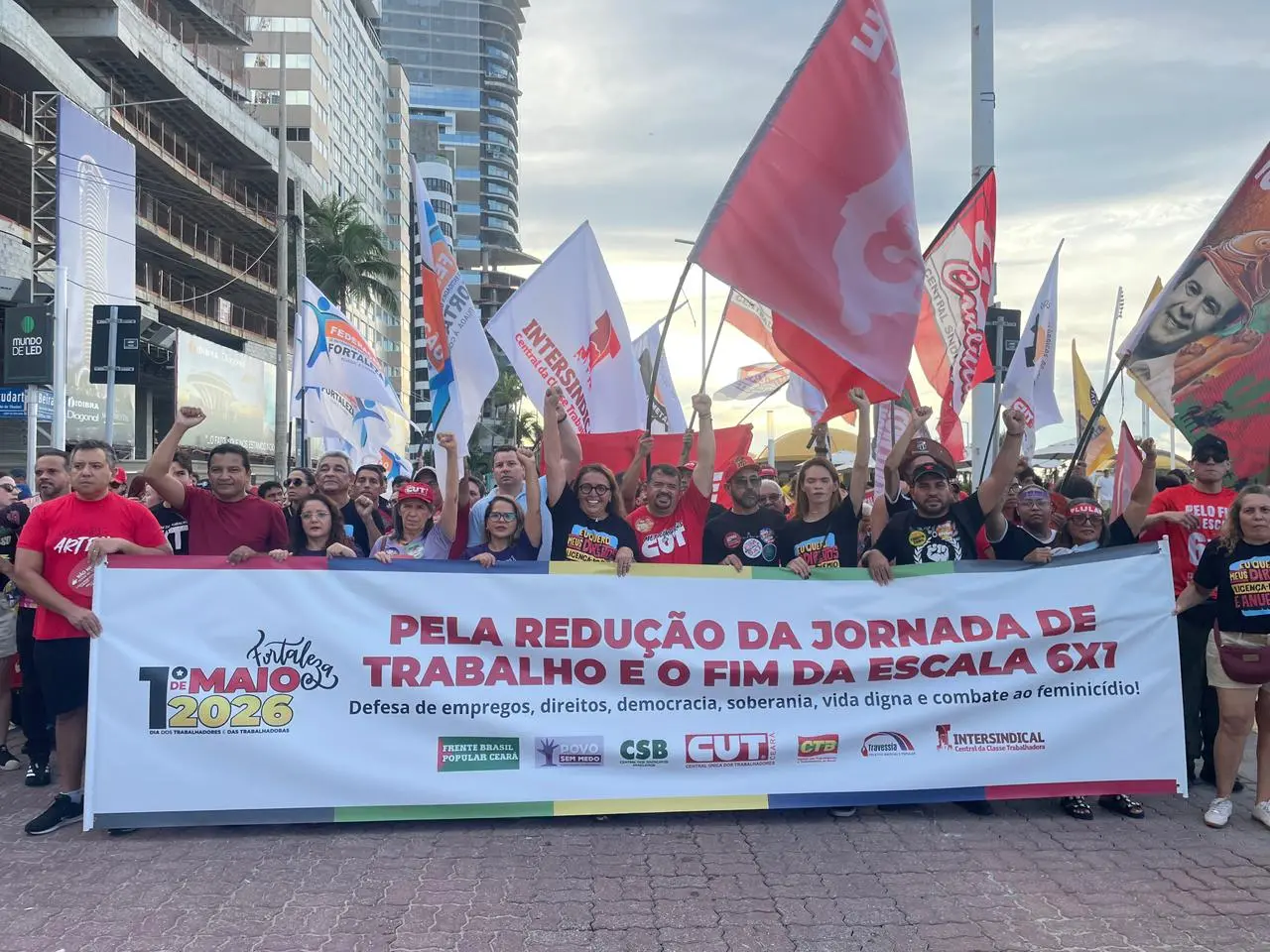 Grupo de pessoas com bandeiras segura uma faixa escrito:Pela redução da jornada de trabaho e o fim da escala 6x1.