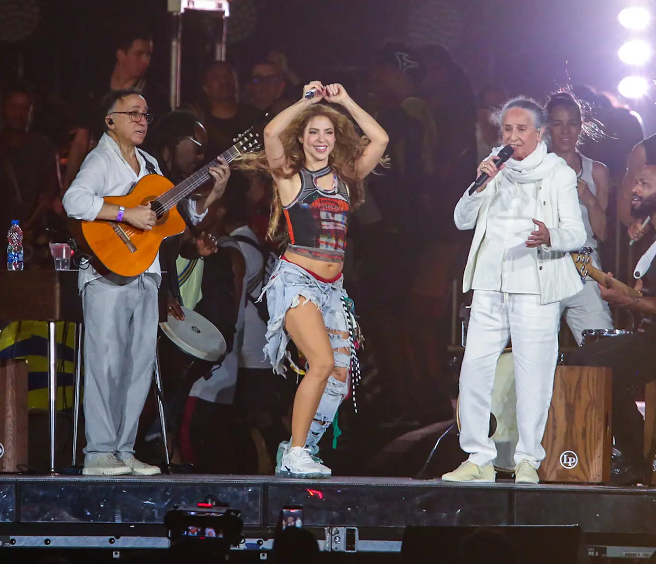 Homem pardo de blusa branca com violão, mulher loira de blusa curta preta e calça jeans e mulher de cabelos brancos com blusa e calça branca performando em palco.