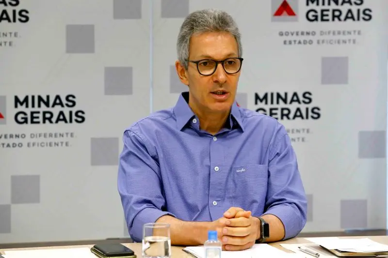 Foto que contém Romeu Zema. Ele está sentado, com óculos de aro grande, e veste uma camisa azul.