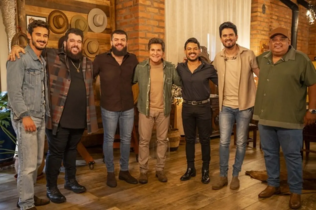 Grupo de seis homens brancos sorrindo em ambiente interno, com decoração de chapéus na parede.