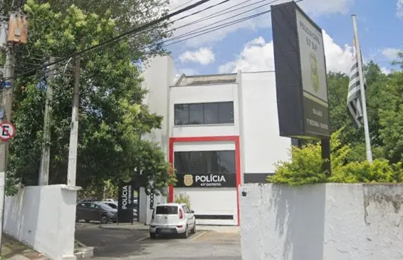 Prédio de delegacia de polícia com fachada branca e detalhes em vermelho, identificado como “Polícia 63º Distrito”. A entrada possui um portão aberto, com um carro branco estacionado em frente. À direita, há uma placa institucional e uma bandeira do estado de São Paulo hasteada. O local é cercado por muros brancos, com árvores e fiação elétrica visíveis ao redor, sob céu parcialmente nublado.