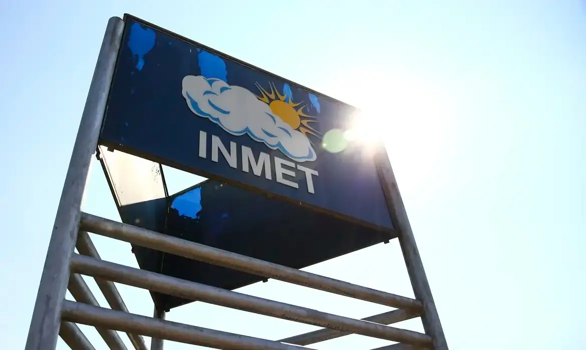 Placa elevada com a sigla “INMET” e ilustração de nuvem e sol, instalada em uma estrutura metálica ao ar livre. A imagem é feita de baixo para cima, com o sol ao fundo provocando reflexo de luz no canto superior, sob céu claro.