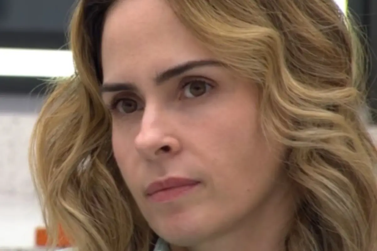 Na imagem, um close-up do rosto de uma mulher com expressão séria e olhar pensativo. Ela tem cabelos loiros ondulados e olhos castanhos. O fundo está desfocado.