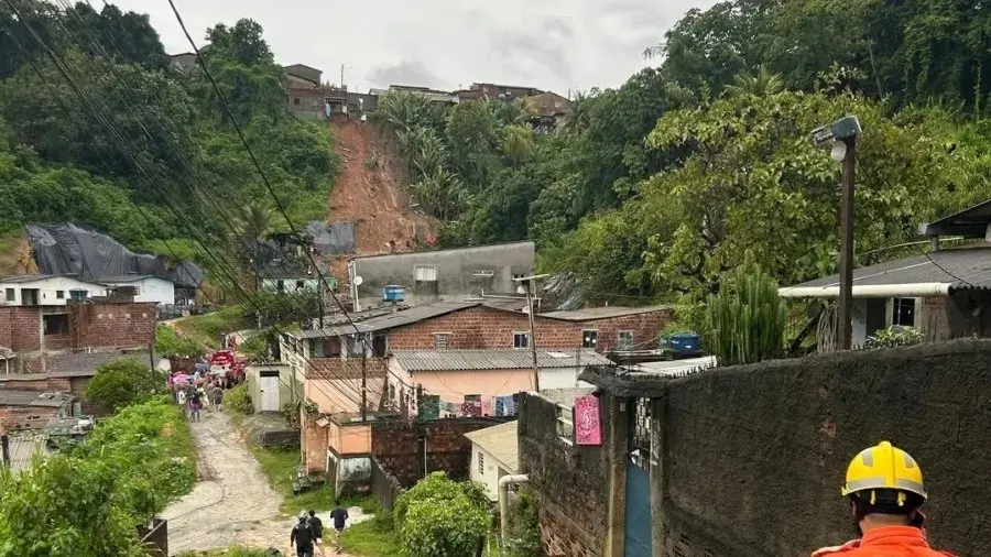Vista de um deslizamento de terra em comunidade de encosta, com casas de diferentes tamanhos, vegetação e trabalhadores de construção na área.