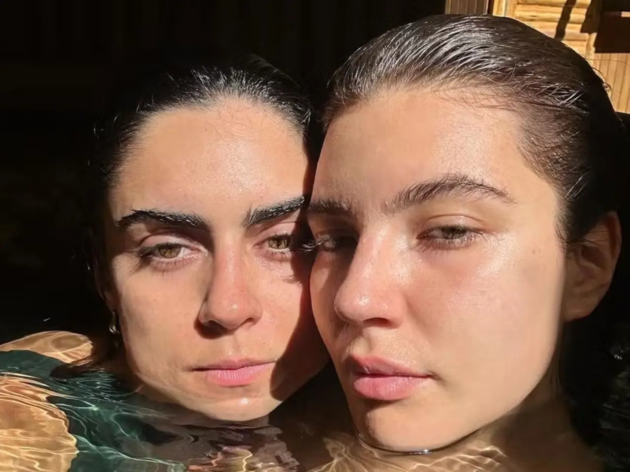 Duas pessoas aparecem parcialmente submersas em água clara, com apenas os rostos visíveis próximos um do outro. A luz do sol incide diretamente sobre as faces, criando reflexos na água e sombras marcadas. O fundo é escuro, contrastando com a iluminação intensa no primeiro plano.