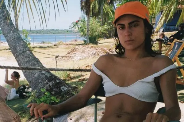 Jovem mulher usando camisa cortada e boné laranja em praia com árvores e água ao fundo, em ambiente ao ar livre com clima ensolarado.