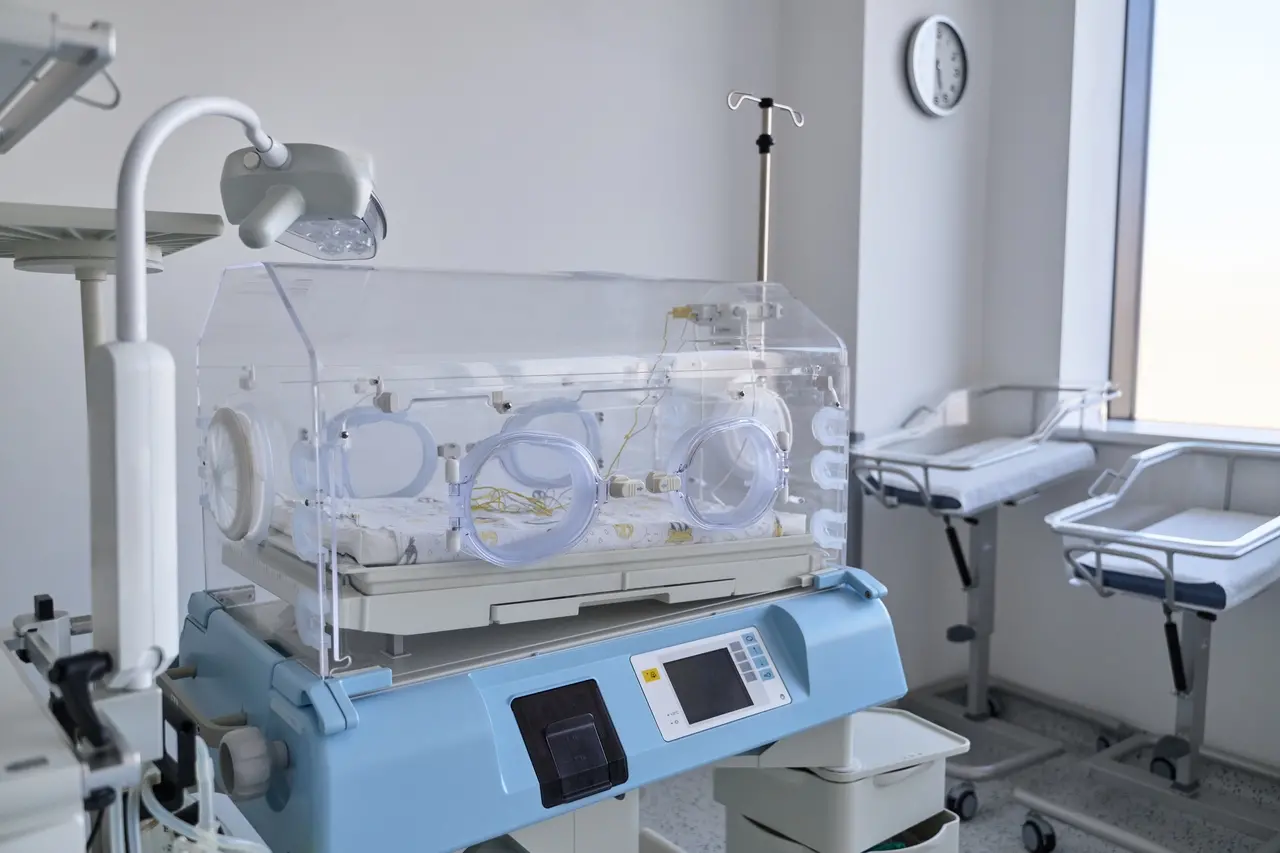 Incubadora neonatal em sala hospitalar, com cúpula transparente, luvas circulares de acesso, monitor digital acoplado e equipamentos médicos ao redor.
