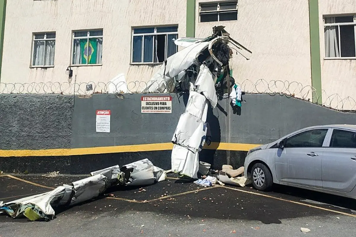 Parte da fuselagem e asas de uma aeronave pequena encostada no muro do prédio e espalhada pelo estacionamento; o carro aparece ao lado e há entulho no chão.