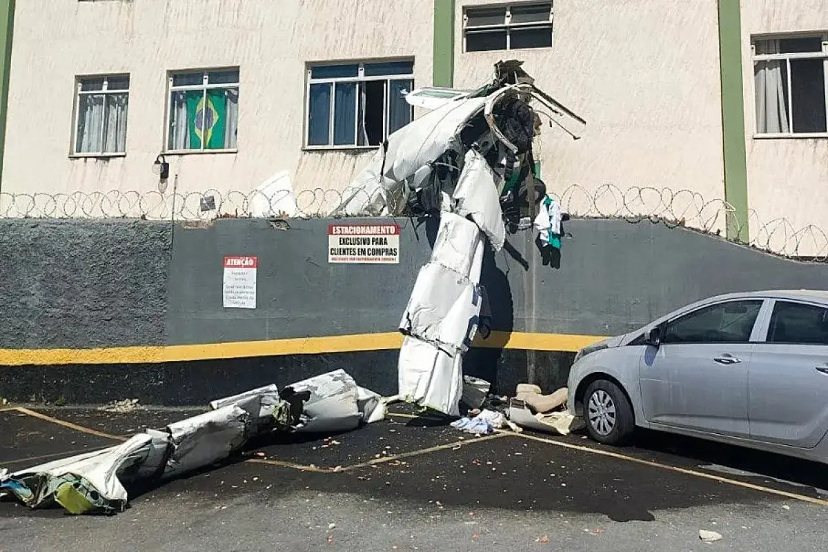 Parte da fuselagem e asas de avião pequeno que caiu e atingiu prédio em BH encostada no muro do prédio e espalhada pelo estacionamento; o carro aparece ao lado e há entulho no chão.