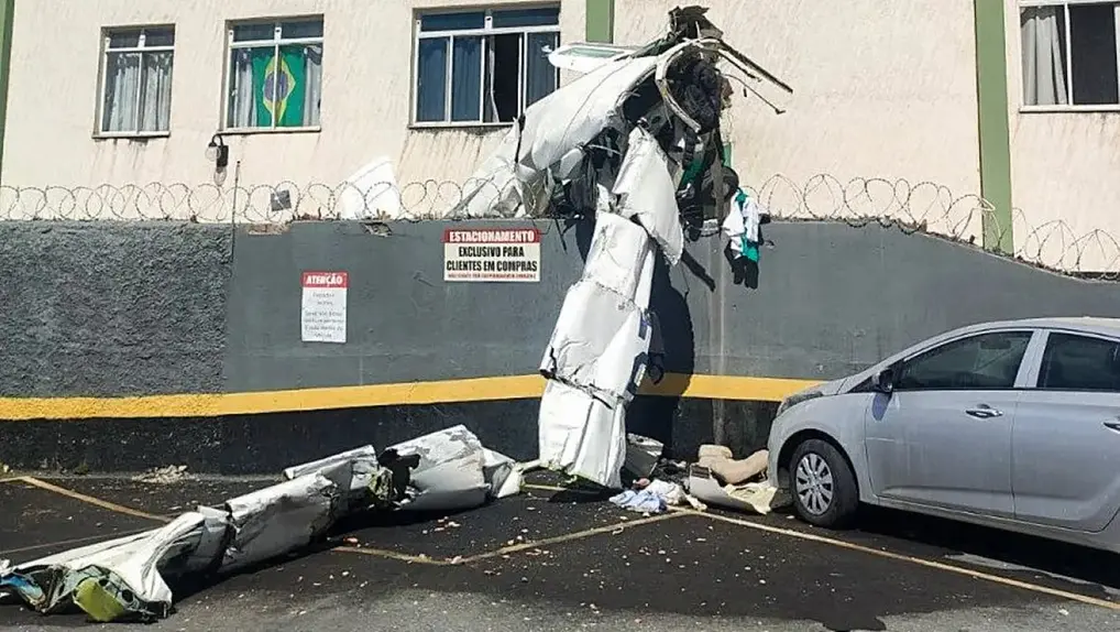 Parte da fuselagem e asas de avião pequeno que caiu e atingiu prédio em BH encostada no muro do prédio e espalhada pelo estacionamento; o carro aparece ao lado e há entulho no chão.