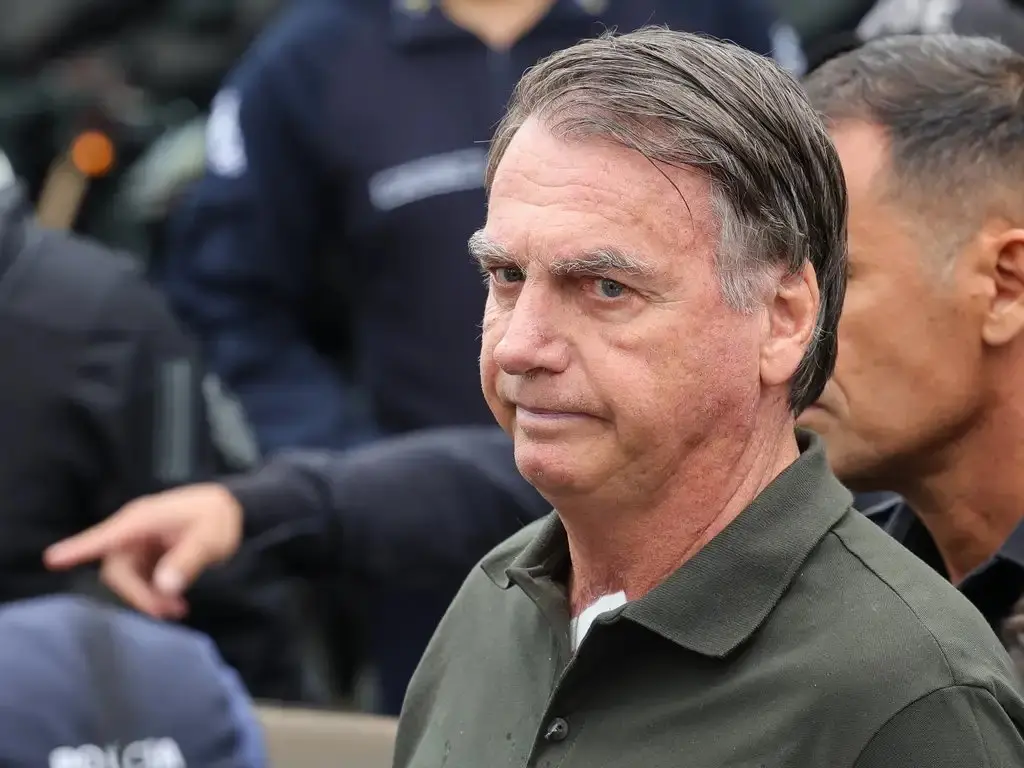 Homem de meia-idade em primeiro plano, usando camisa polo verde, cercado por agentes com coletes escuros ao fundo.