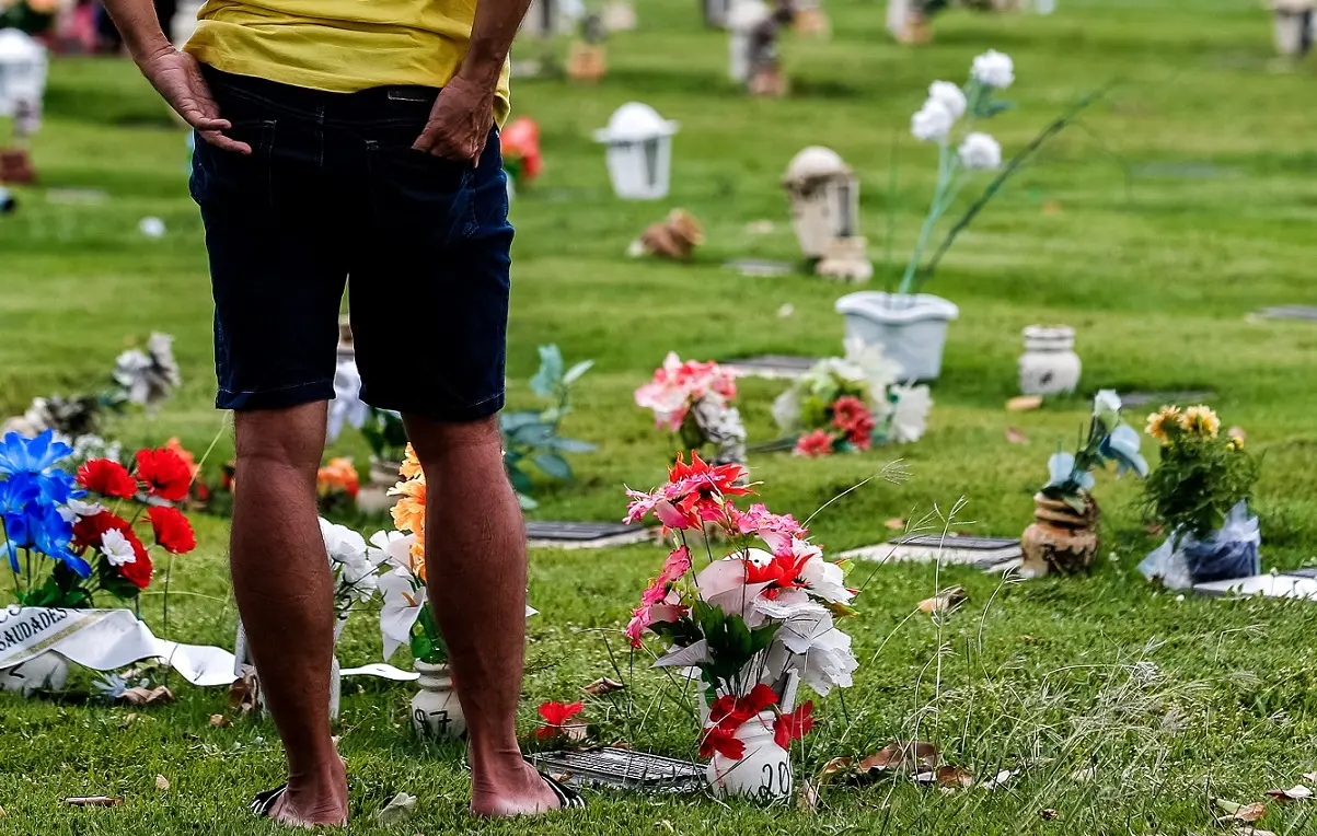 Um homem de costas, vestindo camiseta amarela e bermuda jeans, observa um gramado repleto de flores coloridas deixadas sobre túmulos rasos.