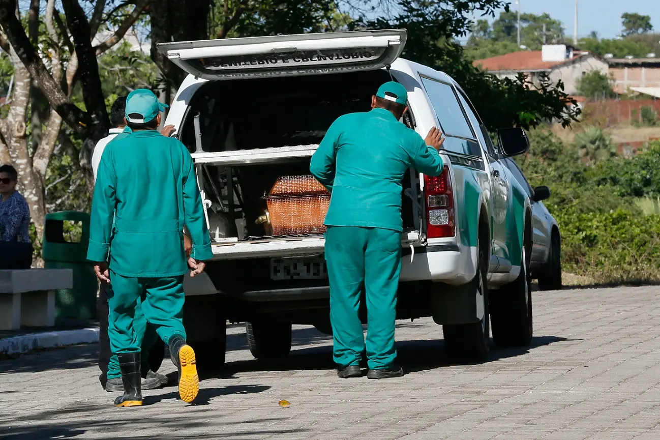 Dois funcionários uniformizados com roupas verdes e bonés trabalham junto à traseira aberta de uma caminhonete branca, onde se vê um caixão de madeira pronto para ser transportado.