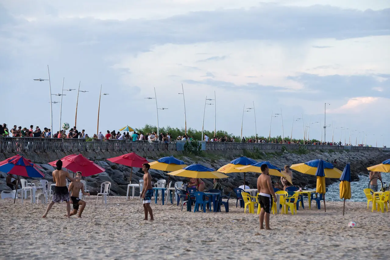 Praia movimentada com pessoas jogando bola na areia e outras sentadas sob guarda-sóis coloridos. Ao fundo, calçadão com grande fluxo de pedestres se estende sobre pedras à beira-mar, sob céu parcialmente nublado.