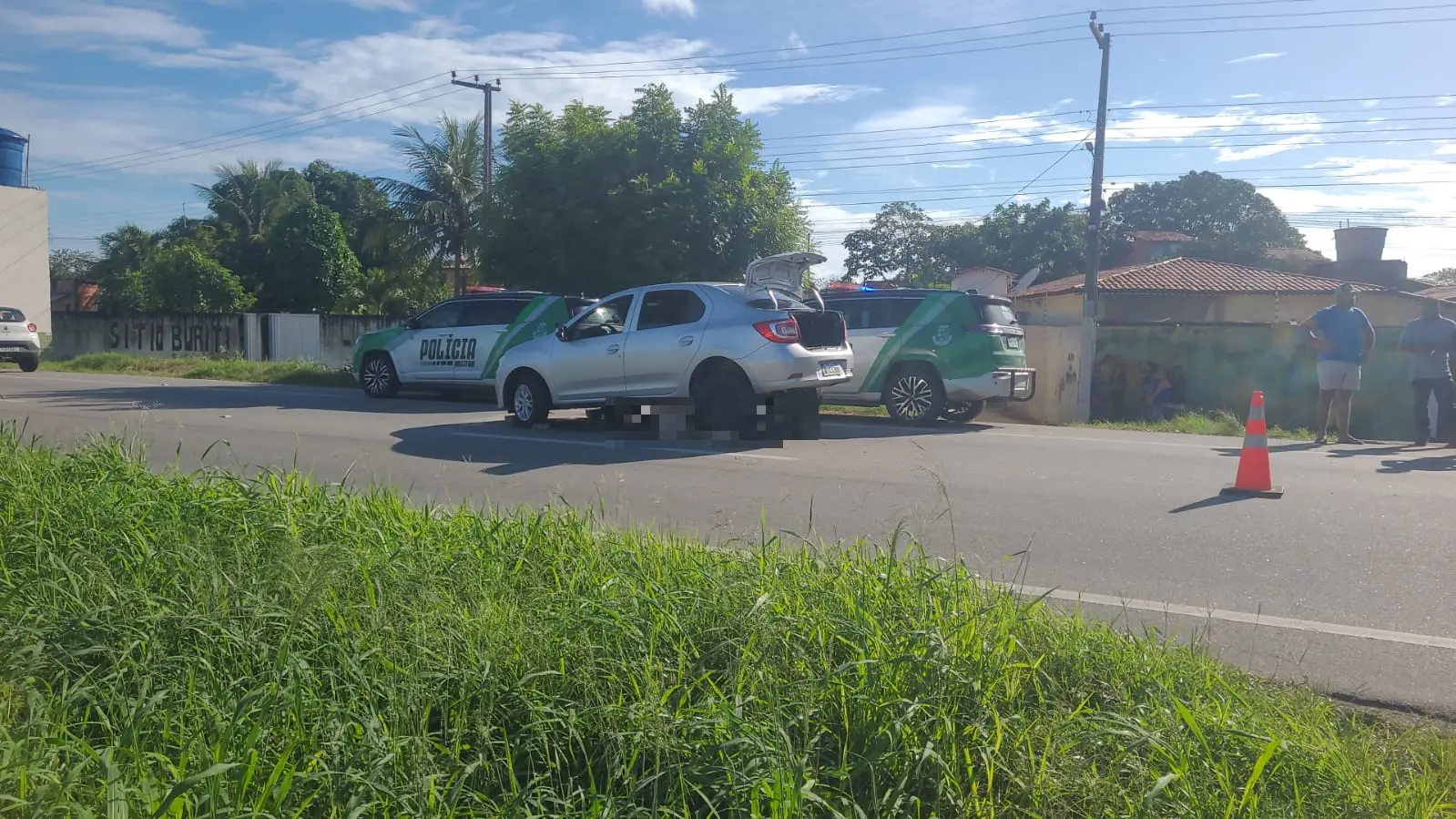 Acidente de trânsito envolvendo dois carros na rua, com uma viatura policial no local e algumas pessoas observando a cena, sob céu ensolarado.