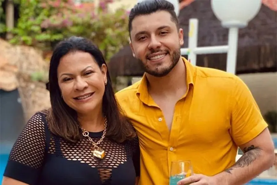 Foto de uma mulher e um homem sorridentes em celebração ao ar livre, com árvores e iluminação ambiente ao fundo, ambos usando roupas casuais e alegres.