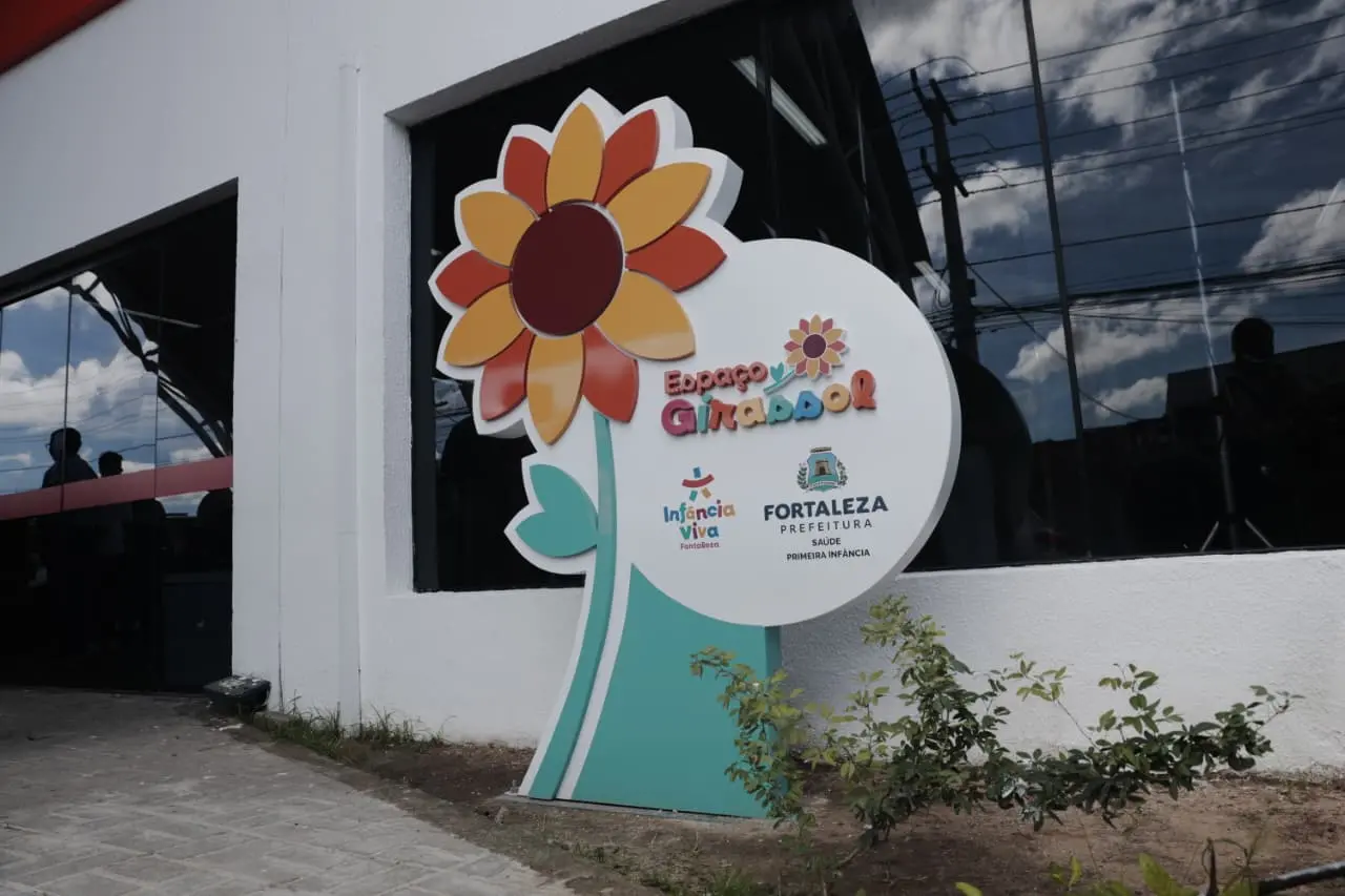Fachada branca com uma placa colorida em formato de girassol gigante. Nela, lê-se Espaço Girassol, acompanhado das logomarcas da Prefeitura de Fortaleza e do programa Infância Viva.