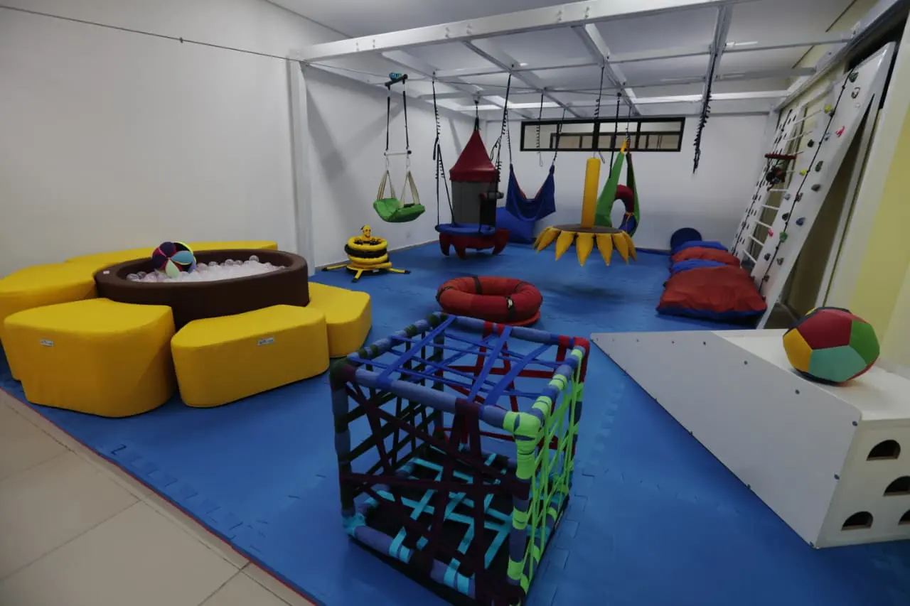 Sala de integração sensorial com piso emborrachado azul e diversos equipamentos lúdicos, incluindo uma piscina de bolinhas amarela em formato de flor, balanços suspensos e uma pequena parede de escalada.