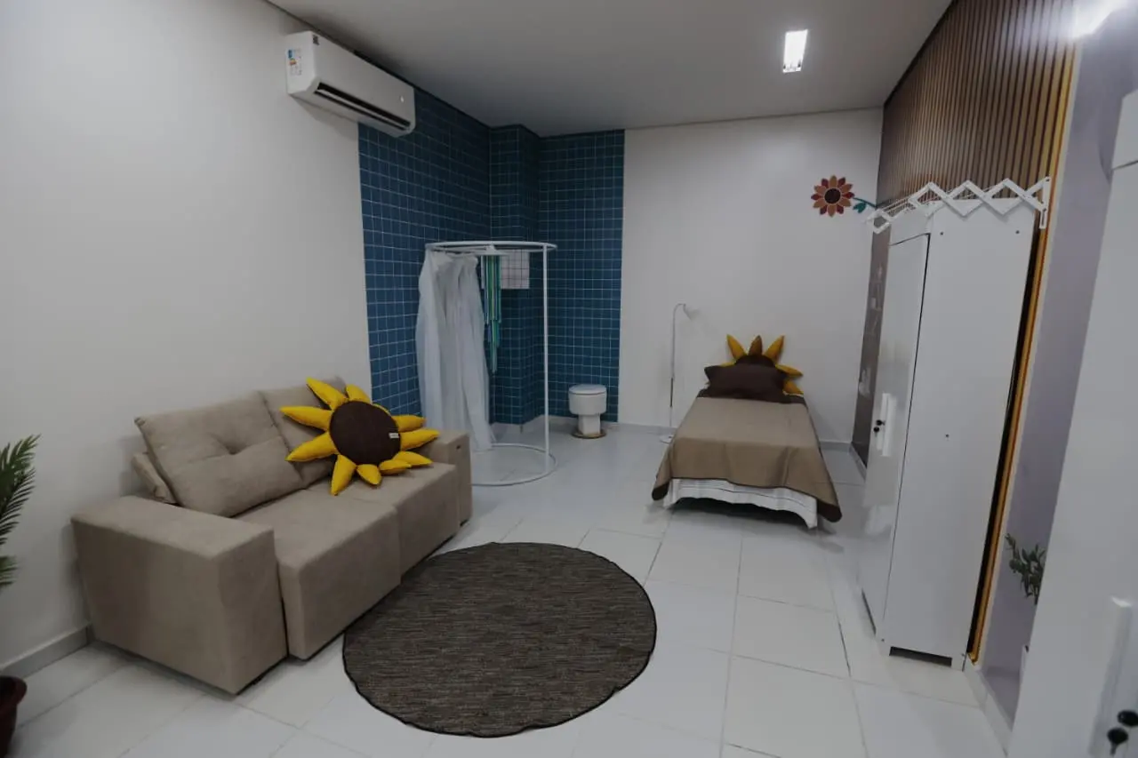 Quarto de acolhimento com paredes brancas e detalhes em azul. Possui sofá bege com almofada de girassol, cama de solteiro, tapete redondo, armário branco e ar-condicionado.