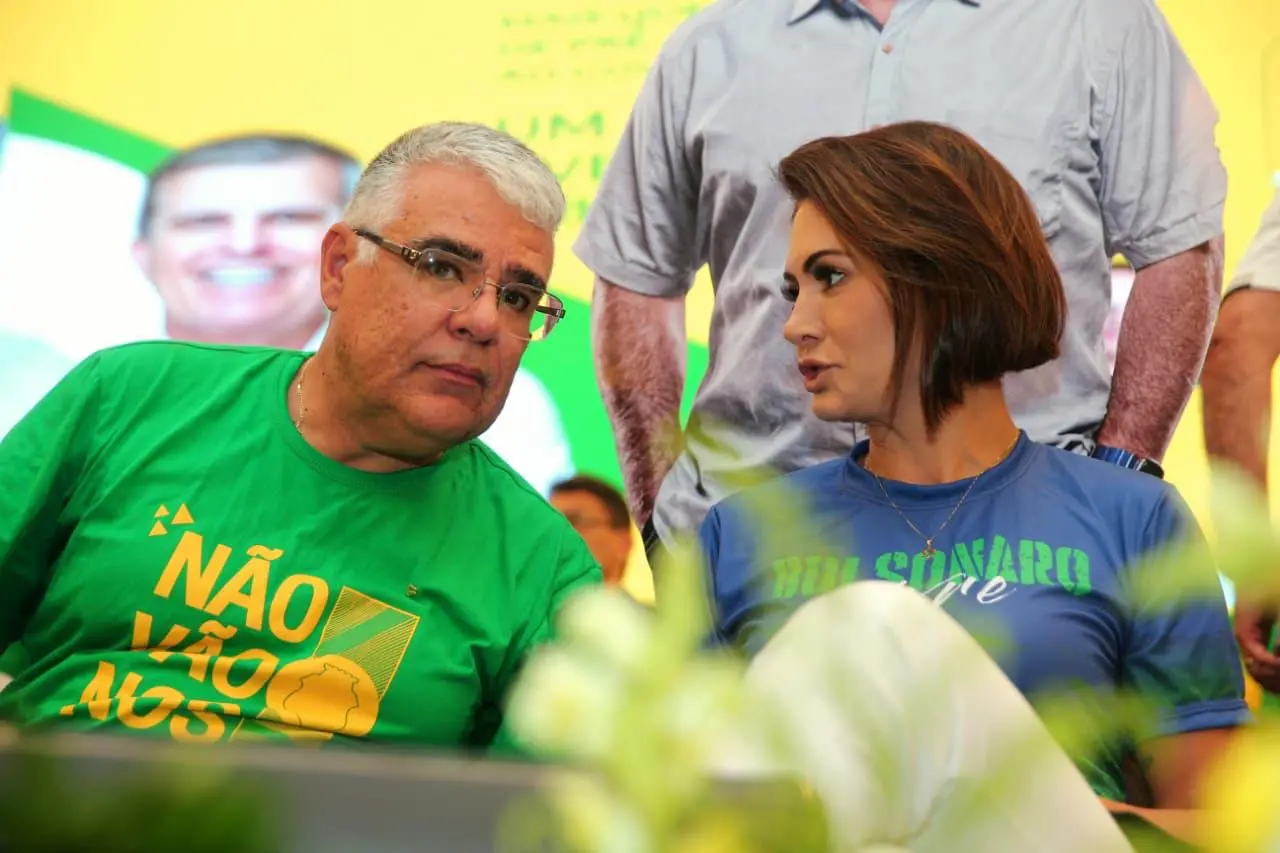 Homem e mulher sentados em evento político conversam entre si; ele usa camiseta verde com a frase “Não vão nos calar”, e ela veste camiseta azul com menção a Bolsonaro, diante de painel com cores verde e amarelo ao fundo.