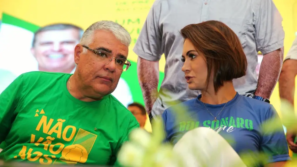 Homem e mulher sentados em evento político conversam entre si; ele usa camiseta verde com a frase “Não vão nos calar”, e ela veste camiseta azul com menção a Bolsonaro, diante de painel com cores verde e amarelo ao fundo.